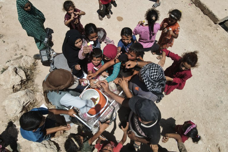 Syrian children gather to buy icecream from an ambulant vendor at a camp for internally displaced people (IDP) near Sarmada, in the northern Syrian province Idlib on June 28, 2024. - After 13 years of civil conflict, lack of international funding has severely undercut the provision of basic services such as water, waste disposal and sanitation in displacement camps in northwest Syria, according to the United Nations. More than five million people, most of them displaced, live in areas outside government control in Syria's north and northwest, according to the UN, many relying on aid to survive. (Photo by AAREF WATAD / AFP)